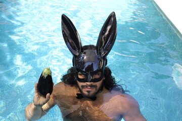 Seductive shirtless muscular man wearing bunny mask in swimming pool holding an eggplant