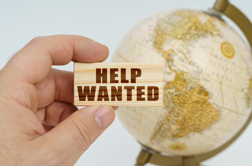 A man holds in his hand a wooden plate on which it is written - HELP WANTED. In the background is a globe.