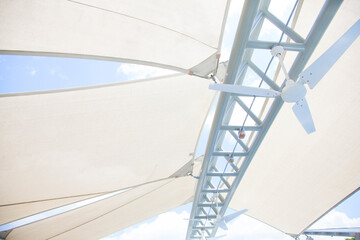 Looking up at a bright white outdoor canvas canopy and geometric architectural strut with a ceiling fan, protecting patio from the summer sun and bright blue sky - obrazy, fototapety, plakaty
