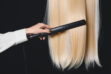 Woman straightening hair with flat iron on dark background