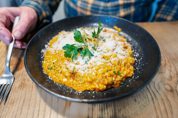 Risotto in a large dish with parmesan cheese. A man is sitting at at rustic wooden table holding a fork.
