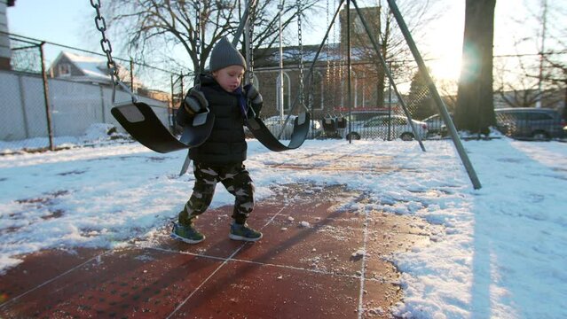 Adorable 5 years old caucasian boy is dancing on slippery playground surface in a funny way holding two swings. Winter in NYC, USA