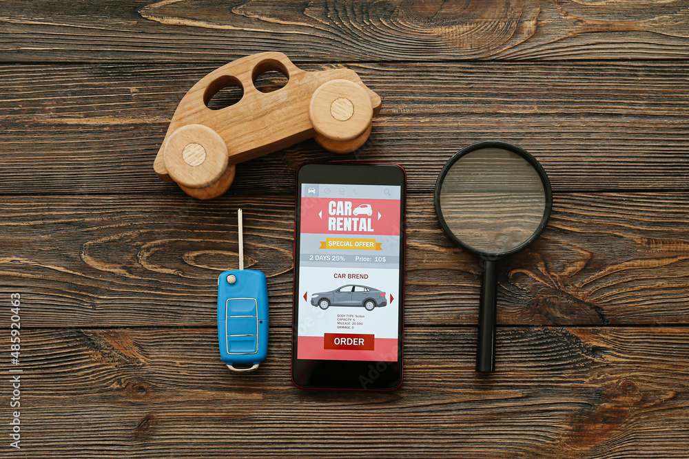 Wall mural Mobile phone with open car rent app, toy, key and magnifier on dark wooden background