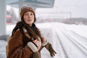 Young trendy asian woman in brown sheepskin coat waiting for train at railroad station in snowy weather. Female tourist in winter coat wait on railway platform for train to arrive. Public transport.