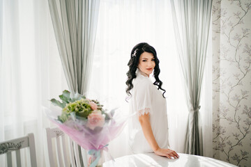 Fototapeta premium pregnant bride brunette in white pajamas by window and table with bouquet. 