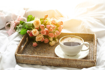 Tray with cup of coffee and bouquet of roses on soft bed