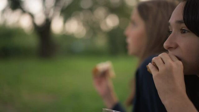 Side view teenage girl on the right eating sandwich with blurred friend at background. Positive relaxed Caucasian teenagers enjoying picnic at beautiful landscape. Mockup template. Slow motion