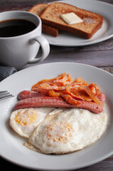 side view vertical image of well done fried eggs with sausage and tomato and onion spicy salsa on a white plate with a fork on the side, a cup of black coffee with toasted bread  and butter.