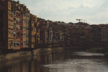 Dark looking panorama of Girona from the river, looking away from Girona cathedral of Saint Mary and red Eiffel bridge. Greenery on the water is seen.