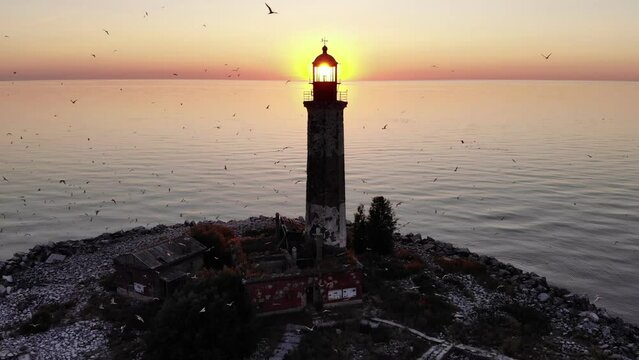 Bright setting sun shines directly through lantern room at top of lighthouse, alarmed seagulls fly around. Dramatic aerial view of an old but functioning lighthouse on small artificial island "Sukho"