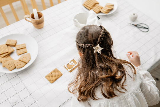 Adorable brown-haired girl wearing white blose decorate with icing gingerbread houses at the kitchen. Christmas sweets, popular Christmas decorations. Casual lifestyle photo in real life interior