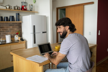 Young business man working at home in his kitchen with laptop and papers on kitchen wooden desk. Home office concept.