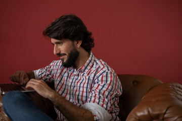 Obraz premium Young business man working at home with laptop on a brown arm chair with a red painted wall in the background. Home office concept.