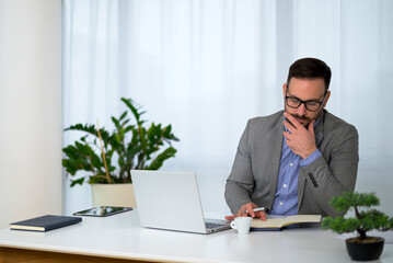 Fototapeta premium serious young businessman with short stylish beard and glasses sitting at office and taking notes