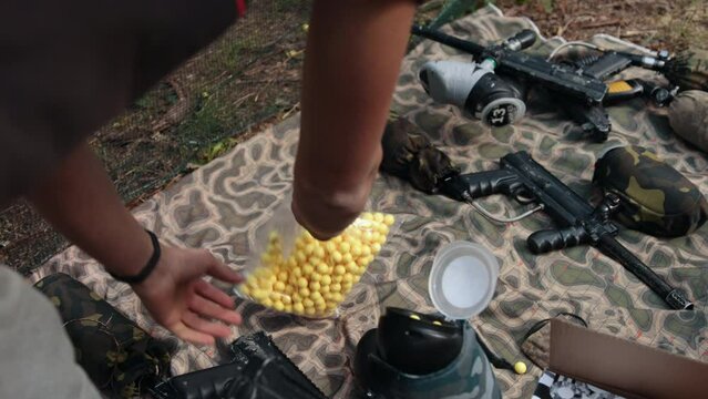 Men's hands pour yellow paintball balls into the weapon tank. Preparing for the game. Close-up. There are rifles nearby. Outdoor activities. Imitation of war
