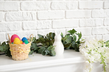 Basket with Easter eggs, decor, eucalyptus and flowers on mantelpiece near white brick wall