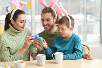 Happy parents with their little son in bunny ears breaking Easter eggs at table