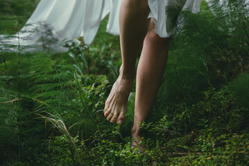 young girl goes barefoot in a white dress on the green grass in the woods on a sunny day, front view close up