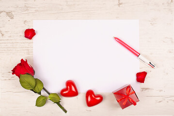A red rose, two decorative hearts and a gift box lie on a table with a white sheet of paper and pen. Mockup for writing text congratulations on Valentine's Day or declaration of love, or wedding,