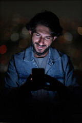 Front view of young man using a smartphone at night time with city view landscape in the background. High quality photo