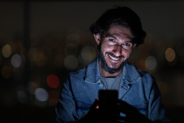 Front view of young man using a smartphone at night time with city view landscape in the background. High quality photo