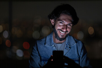 Front view of young man using a smartphone at night time with city view landscape in the background. High quality photo