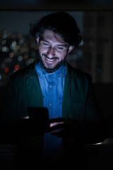 Front view of young man using a smartphone at night time with city view landscape in the background. High quality photo