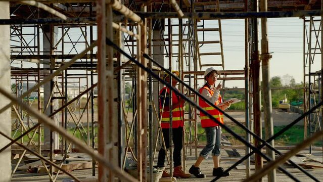 young Asian female engineer and foreman with blueprints is walking to check whether the construction works according to the plan or not at the construction site.
