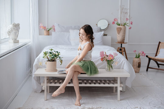 A beautiful little girl with curly hair in a green puffy skirt sits in a spacious bright room among the flowers. Delicate photo. The child poses in the bedroom by the big white bed. 