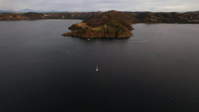 Flying drone approaching single sailboat navigating in parallel to Costa Rica rocky coastline