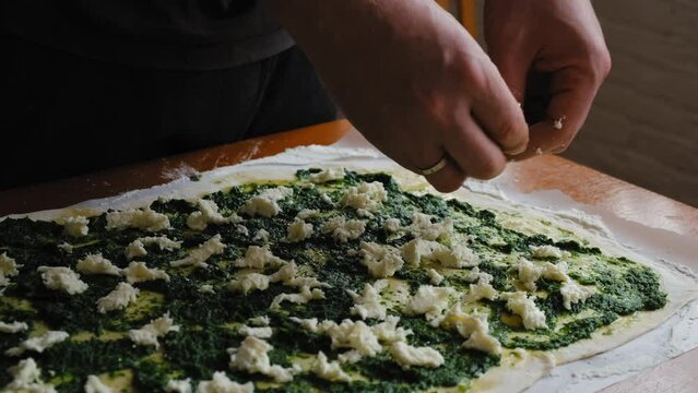 Man cooks a Viennese strudel with spinach and cheese. Male hands put cheese on raw dough. Home cooking, homemade pastry concept.