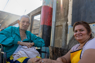 Adult Latin woman with her elderly father in a wheelchair on the outskirts of a poor neighborhood in Latin America
