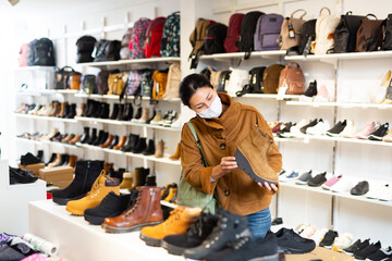 Asian woman in a protective mask, who came to a shoe store for shopping during the pandemic, chooses autumn halfboots ..with high soles