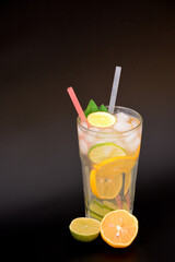 A tall glass of citrus lemonade with ice on a black background next to a sliced ​​lemon and lime.