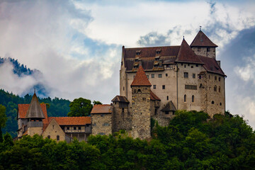 Stone fortified Gutenberg Castle among green trees on top of hill on background with mountains and cloudy sky, Balzers, Liechtenstein