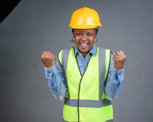 Studio portrait of a happy African Nigerian career lady or female engineer wearing a yellow safety helmet and a reflective jacket and raising her fits up in jubilation