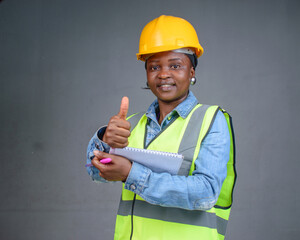 Studio portrait of a happy African Nigerian career lady or female engineer wearing a yellow safety helmet, reflective jacket and having a pen and writing pad in her hands while doing thumbs up
