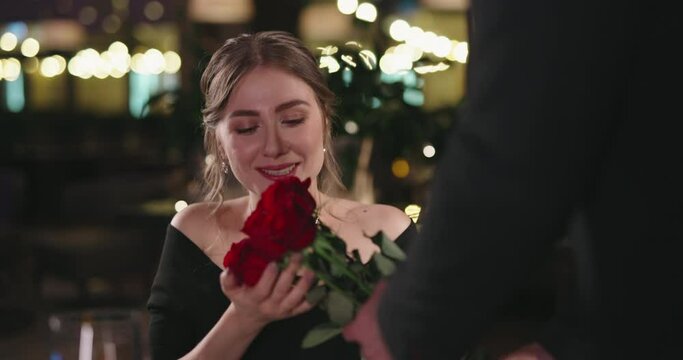 Man in suit approaching beautiful caucasian girl while holding bouquet of red roses behind the back. Guy surprising girlfriend before date 4k footage