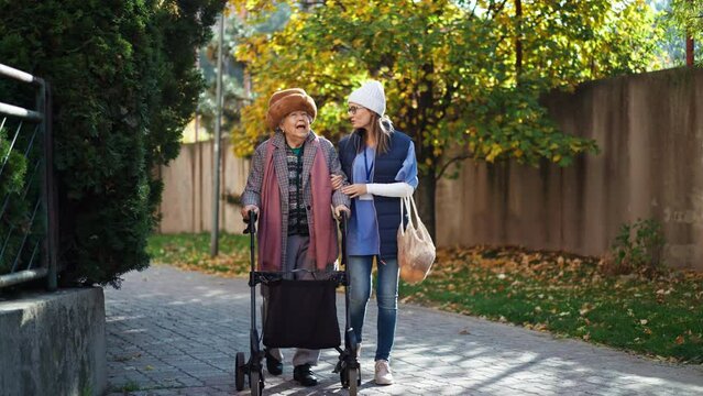Senior woman and caregiver outdoors on a walk with walker in town, talking.