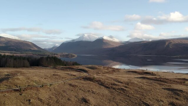 Aerial drone footage flying forward and arising above bracken and moorland to reveal Glen Etive and Loch Etive in Scotland in the morning light, with snow-capped mountains, a forest and still water