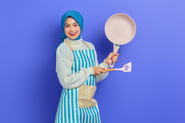 Portrait of cheerful young Asian Muslim woman housewife wearing apron showing frying pan and pointing to copy space with spatula isolated on purple background. Housekeeping concept