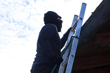 thief with balaclava and crowbar on a ladder trying to sneak into a house