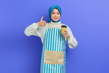 Smiling young Asian muslim woman in her 20s wearing hijab and apron holding paper coffee cup while showing thumb up isolated over purple background. People housewife muslim lifestyle concept