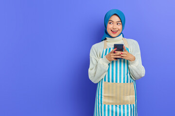 Portrait of smiling young Asian Muslim woman 20s wearing hijab and apron typing sms messages in mobile phone while look aside isolated on purple background. People housewife muslim lifestyle concept