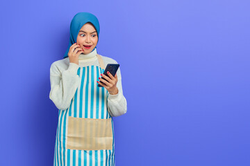 Beautiful young Asian muslim woman wearing hijab and apron,  looking at  ig promo on smartphone with surprised expression isolated over purple background. People housewife muslim lifestyle concept