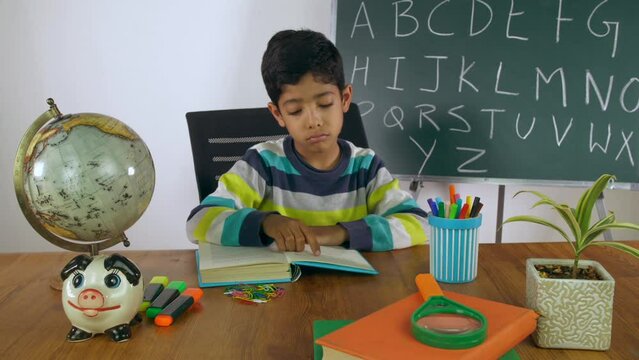 A reluctant and tired schoolchild in casual clothes resting on the desk with a book. Books  globe  marker  and a blackboard together in the classroom - study material  homeschooling  stationery