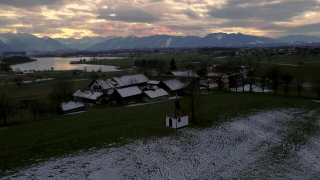 Circling a chapel at scenic Bavarian lake Staffelsee in south Germany close to the alps with a  vibrant dramatic sunset cloud sky and idyllic mountains and snowy countryside fields. 4K aerial drone