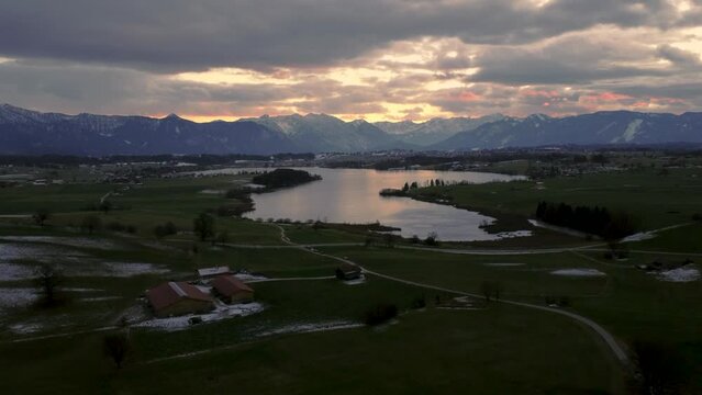 Scenic Bavarian lake Staffelsee in winter in south Germany close to the alps with a  vibrant dramatic sunset cloud sky and idyllic mountains and green countryside fields. 4K UHD aerial drone footage