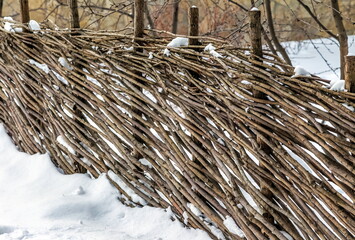 A fence made of tree branches (wattle fence) in close-up against the background of snow and trees in winter