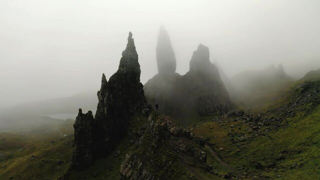 Man in dark clothes stands on top of a crag with in the background the enormously high rock peaks shrouded in fog called Old man of Storr rock in Skye mountains in Scotland. Drone dolly shot